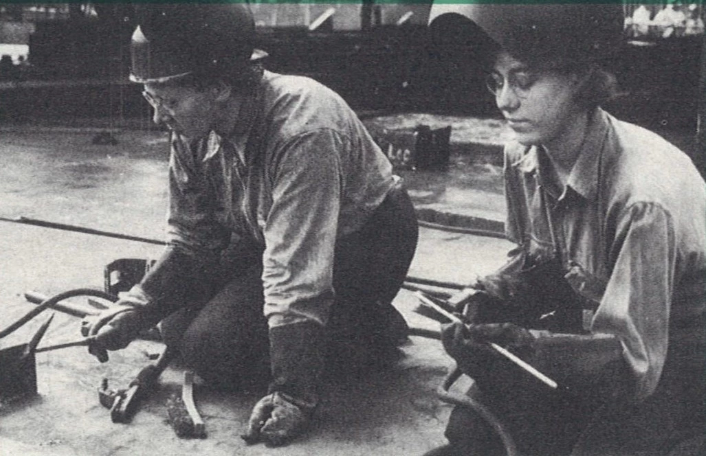 3.023BosnWhistle(1) An image of two women sitting together on a ship floor, wearing helmets and other safety equipment, welding pieces of metal together