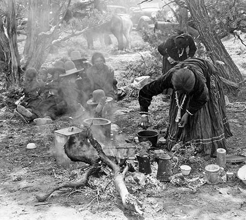 Navajo people harvesting pinons circa 1932 Women bend over harvesting and cooking pinon nuts while there is a crowd of people sitting on the ground in the left background.