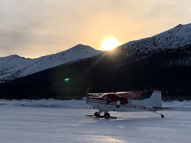 A ski plane sits on a frozen lake with mountains, and a setting sun in the background. 