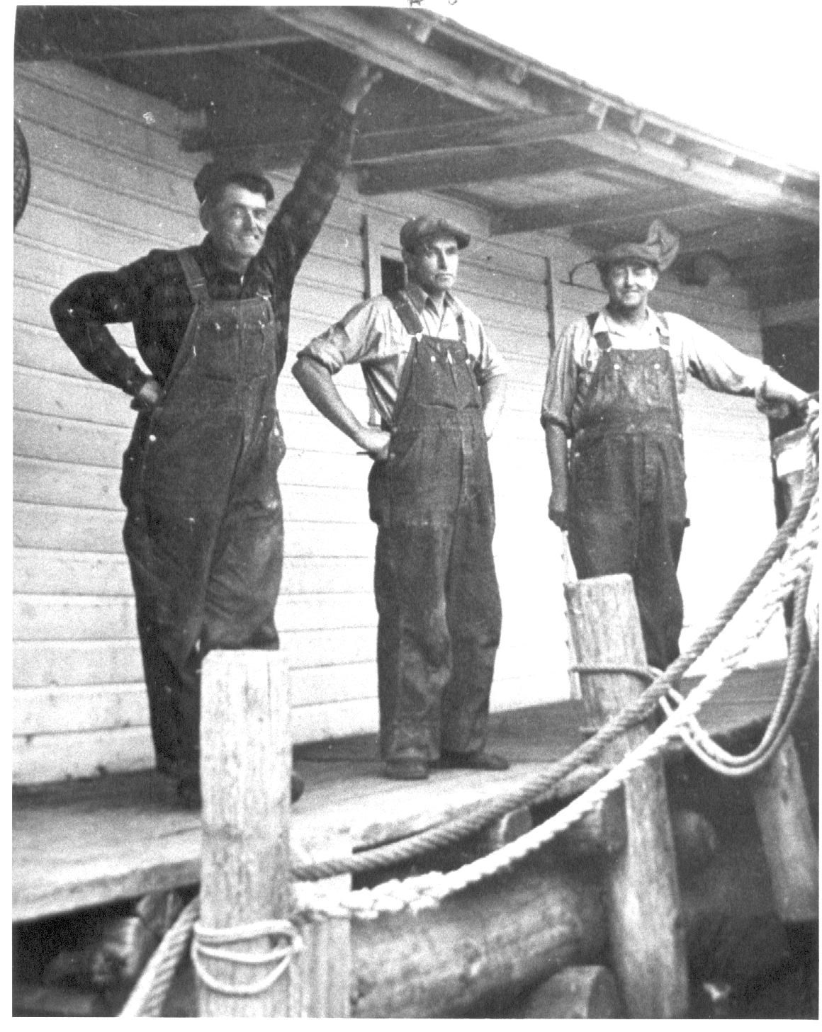 Three men standing under the eave of a building with mooring ropes across the bottom right corned of the photo.