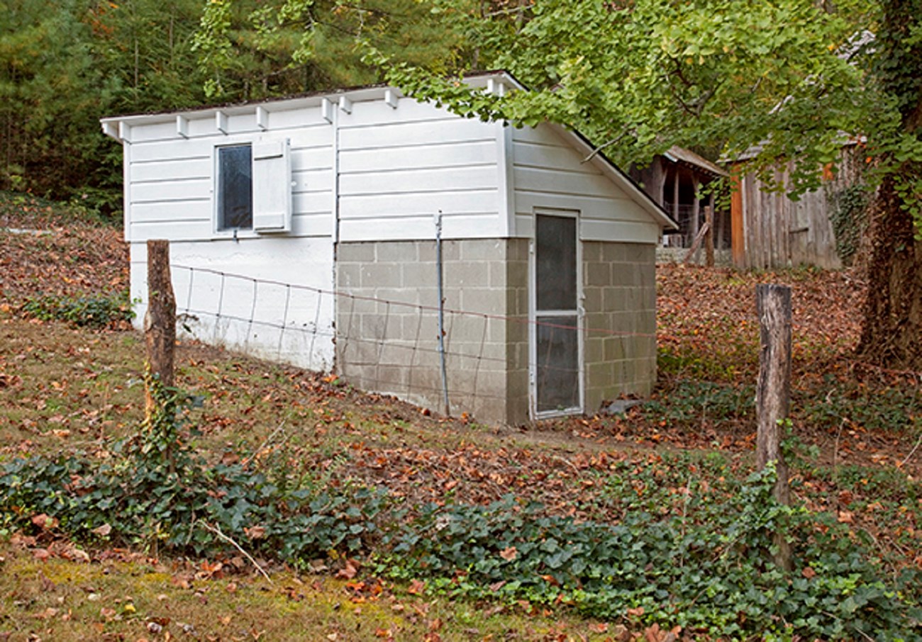 A small wood and block building, with a door on one end and a window on one side, sits behind a wire fence with other buildings in background