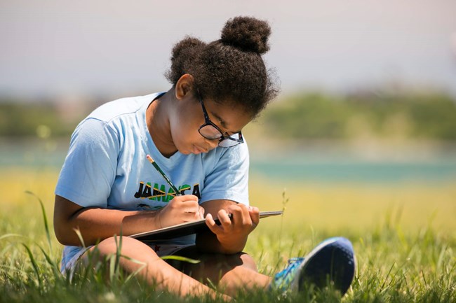 an adolescent sitting on the ground writing in a journal