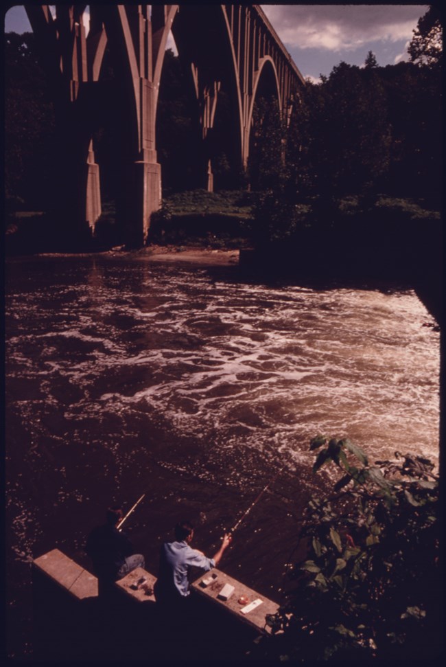 Two fisherman stand along the Cuyahoga River, fishing poles in hand