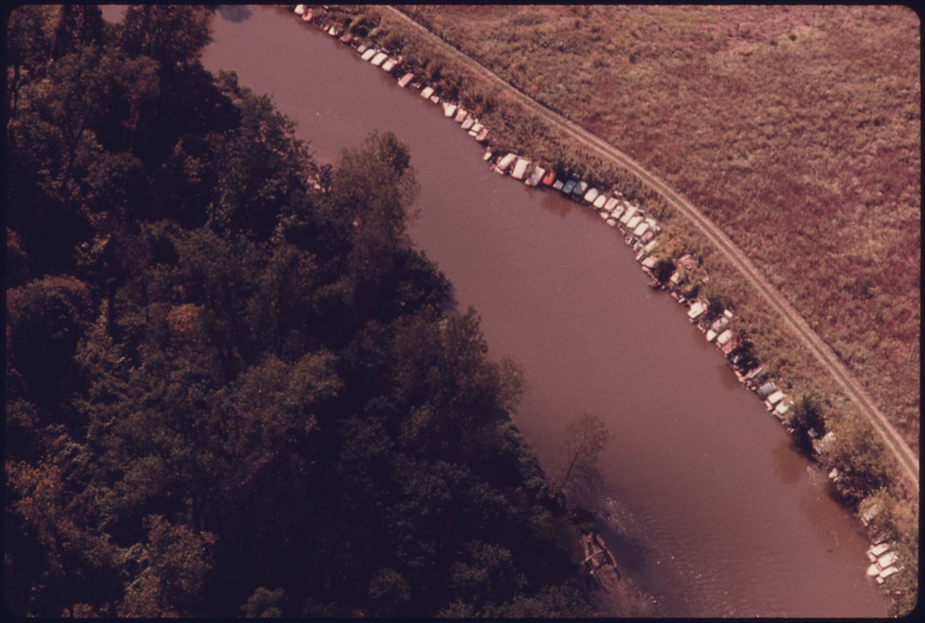 An aerial view shows 50+ old cars lining a riverbank with a field behind and forest opposite.