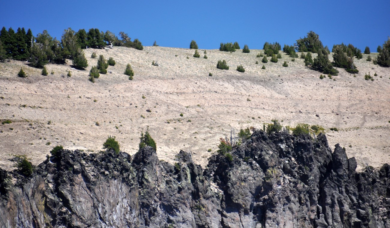 photo of a jagged cliff of lava rock topped by thick beds of fine-grained volcanic ash and pumice