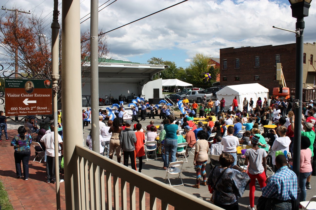 Festival in Jackson Ward Large crowd gathered on a paved area in front of a stage, viewed from the porch of Maggie L. Walker National Historic Site, with a visitor center sign visible in the far left of the frame.