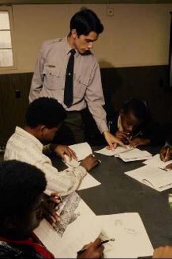 A National Park Service employee talks to children who are participating in the NEED program. All of the children in the photograph are African American, and they are all writing in notebooks and reviewing reading material.