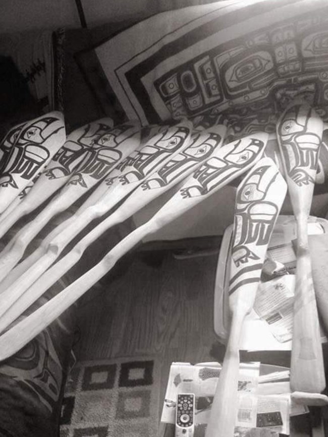 Nine hand carved wooden canoe paddles with Northwest Coast stylized Raven's and Eagles painted on them. A Chilkat blanket with Northwest Coast designs is in the background.