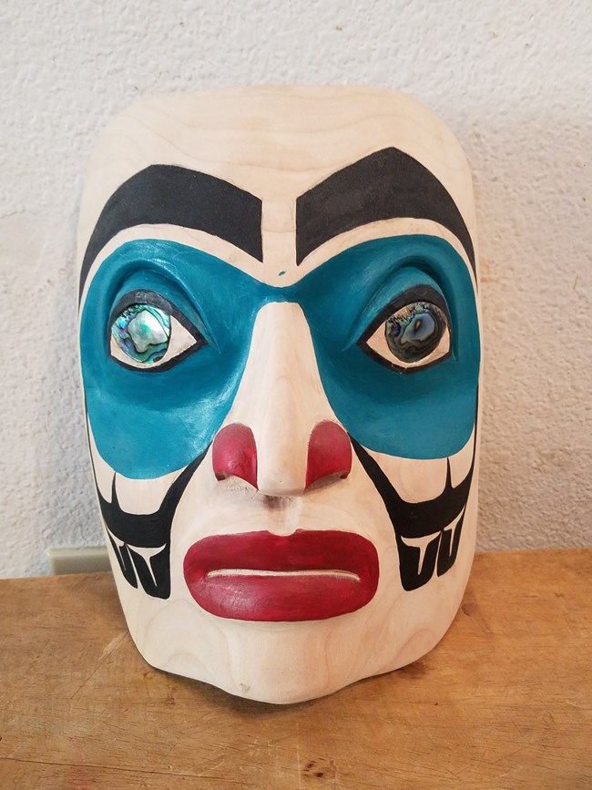 A mask carved from wood in a Northwest Coast Art style. The eyes are inlaid with abalone shells, the area around the eyes is painted blue, the nostrils and mouth are red. The eyebrows and patterns on the cheeks are black.