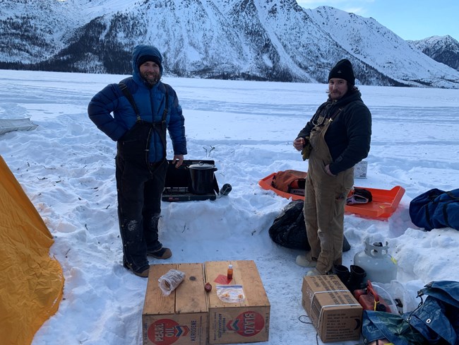 Two men stand near boxes of gear and a tent on a frozen lake. 