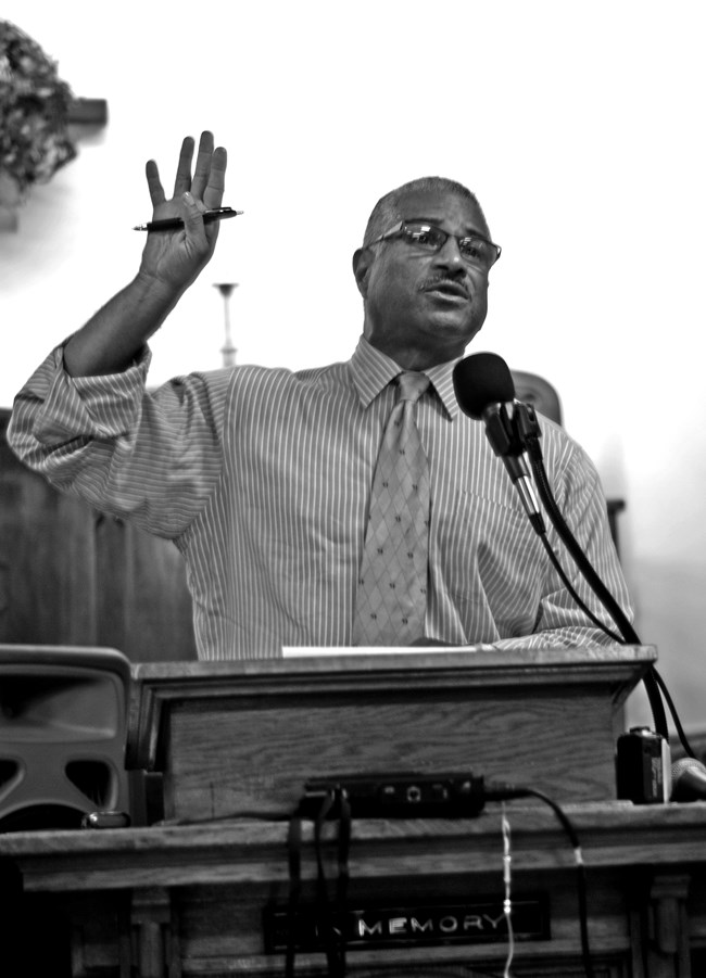 Man stands in front of podium with his arm raised and a pen in his hand.