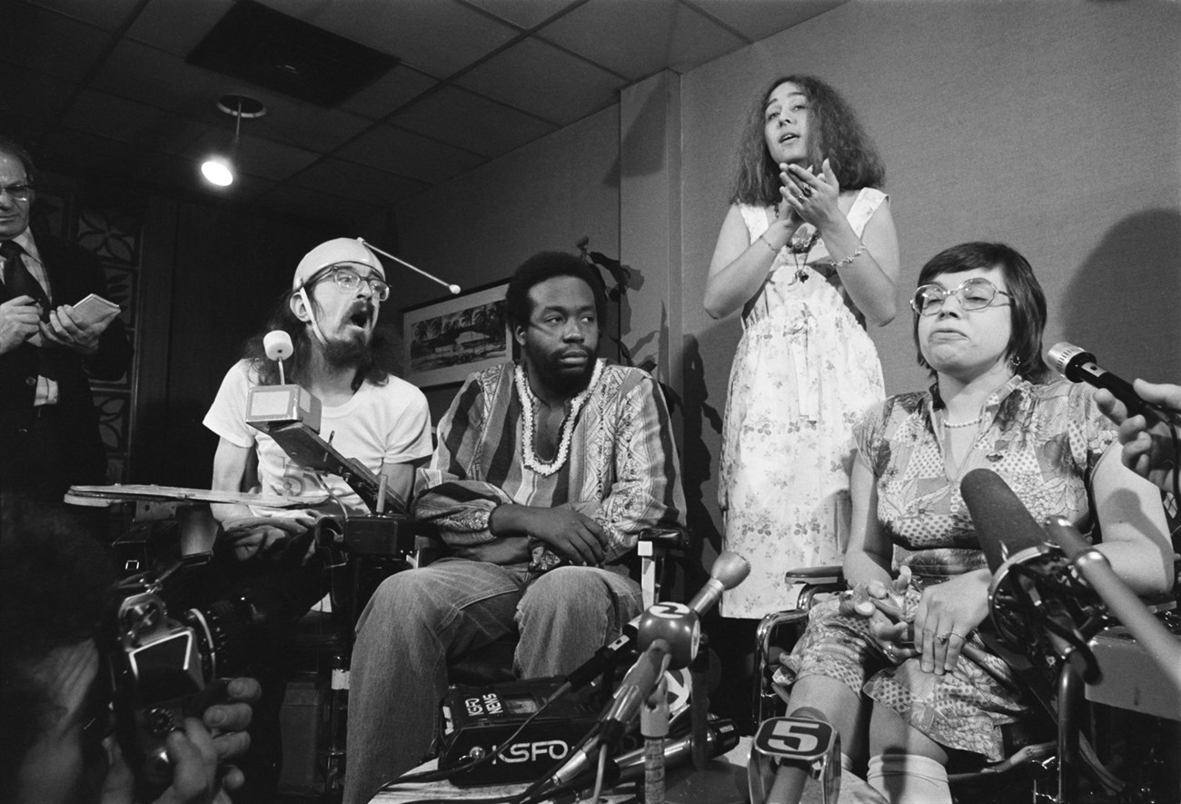 Three people in wheelchairs one white man, one black man, and a white woman speaking into microphones with cameras pointed at them. A white woman stands in the background using American Sign Language.