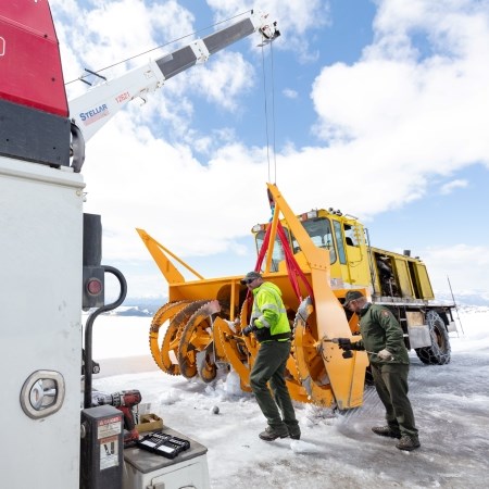 two people repairing a large snowplow on a snowy road