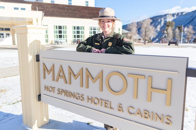 Arrive with Accommodations a park ranger standing behind a sign that reads "Mammoth Hot Springs Hotel"
