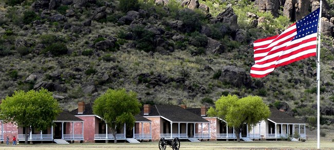 Five stone houses with porches sit on a grassy field.  A rocky ledge is seen in the background.  A cannon and American flag on a flag pole are in the forefront.