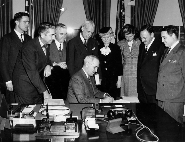 A group stands in a semicircle around President Truman (seated) as he signs a bill.