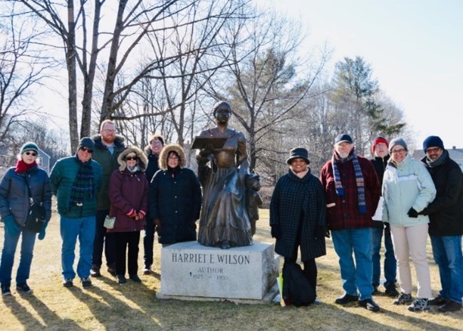 Harriet E. Wilson Memorial. Milford, New Hampshire Bronze memorial statue, with six people standing to its left and five to its right, all in winter garb. Bare trees in background with bright sky shining behind. Stone base of memorial is engraved with text: "Harriet E. Wilson, Author"