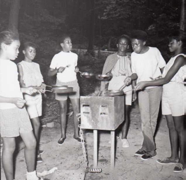 Photograph showing Summer in the Parks participants cooking outdoors on an outdoor grill. All of the participants shown in the photograph are African American women and girls, and they are holding frying pans over the grill.