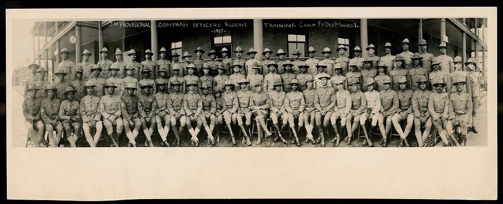 Large group portrait of African American officers wearing military uniforms and broad brimmed hats. Front row is seated with crossed ankles and hands in laps. Middle and back row are standing in front of a building.