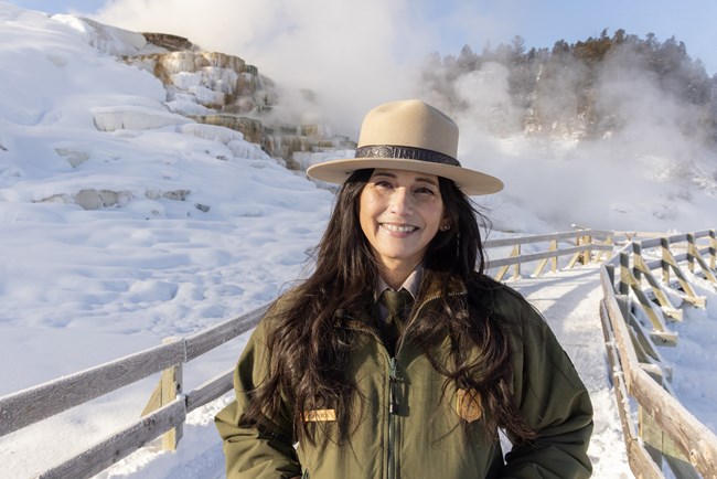 A park ranger smiles on a snow-covered boardwalk in front of a thermal feature.