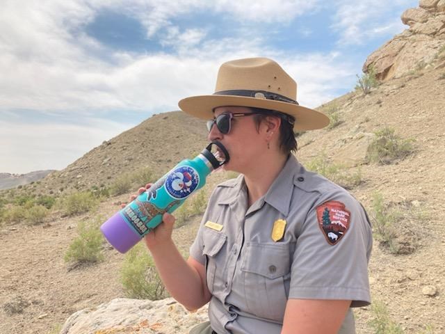 A ranger sits on a rock in front of hillsides, drinking out of a water bottle.
