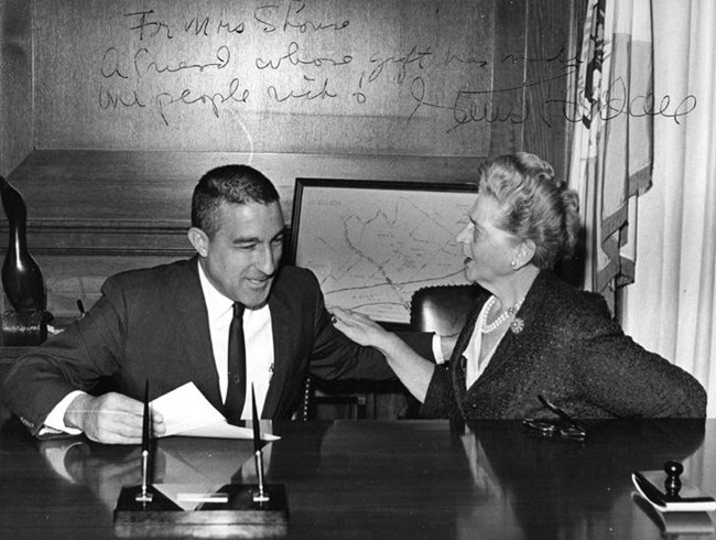Black and white photo of a formally dressed man and woman are seated behind a large desk facing each other having a conversation. A cursive message on the photo reads, "For Mrs. Shouse A Friend whose gift has made the people rich! Stewart L. Udall."