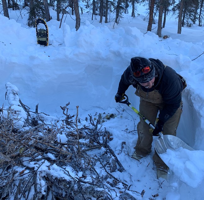 Digging the burn pit at Narvak Lake. A man stands with a shovel in a snowy area with a partially dug pit.