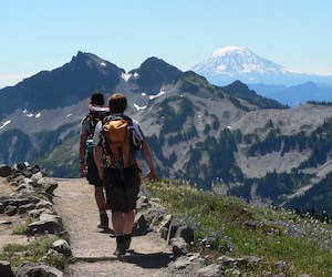 Hikers at Paradise Two hikers with day packs walk along a trail with views of mountain ridges and a distant volcanic peak.