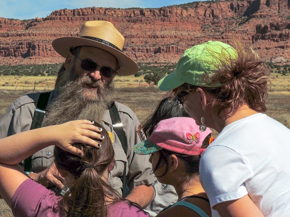 Ranger Ron Brown with Condor Enthusiasts by NPS/Kristen Luetkemeier Park Ranger with a flat hat, dark glasses, and a long beard talks to an adult and children while tall red cliffs tower in the background.