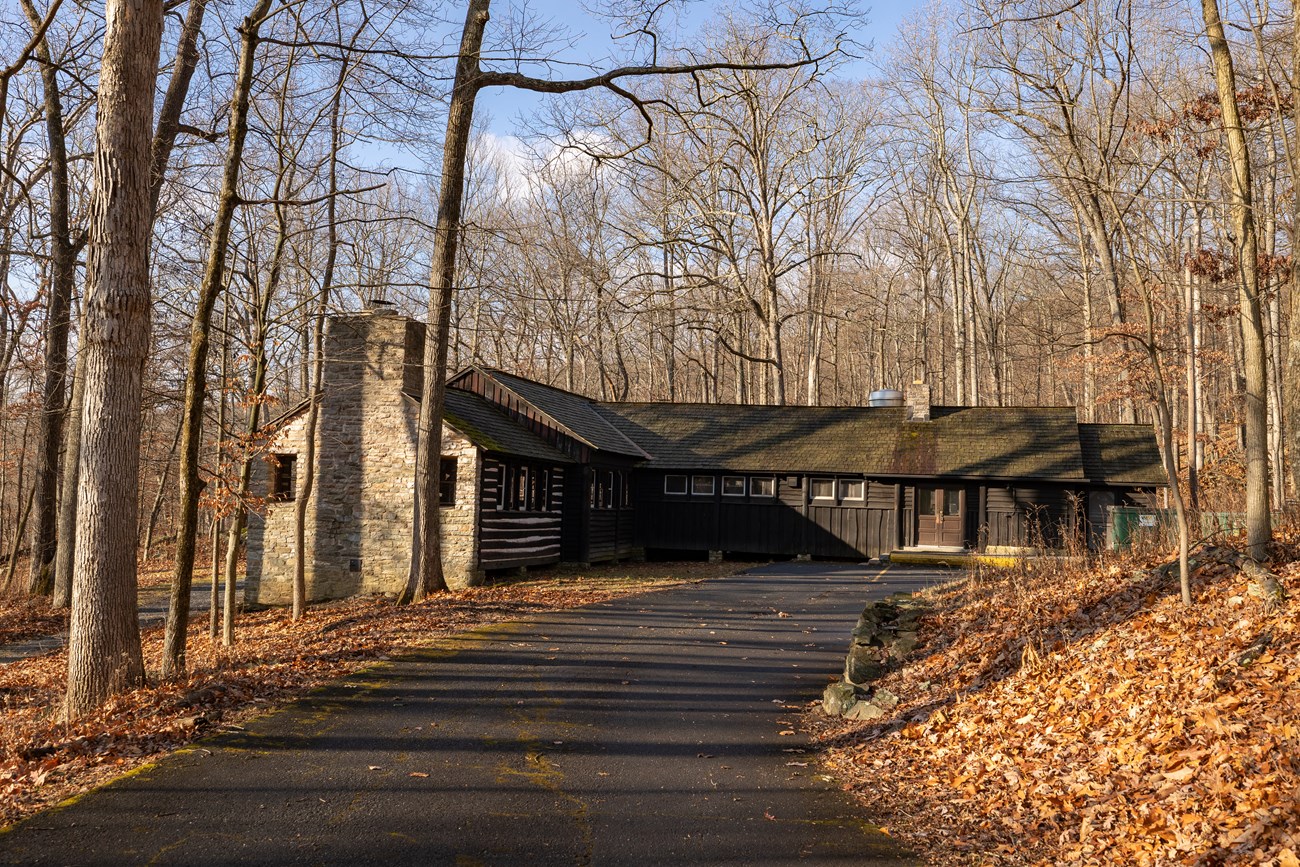 Camp Misty Mount Dining Hall, Catoctin Mountain Park A paved road leads to a large, stone and wood building with two chimneys and a front double door. It's closely surrounded by lots of leafless trees, during what appears to be late fall..
