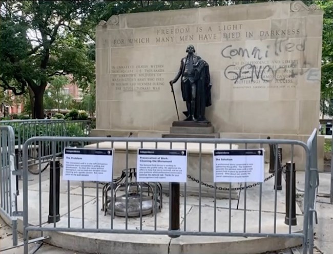 The words "Committed GENOCIDE" spray painted on the monument beside a statue of George Washington. A barrier of metal fences, each with a sign, cordons off the tomb and monument.