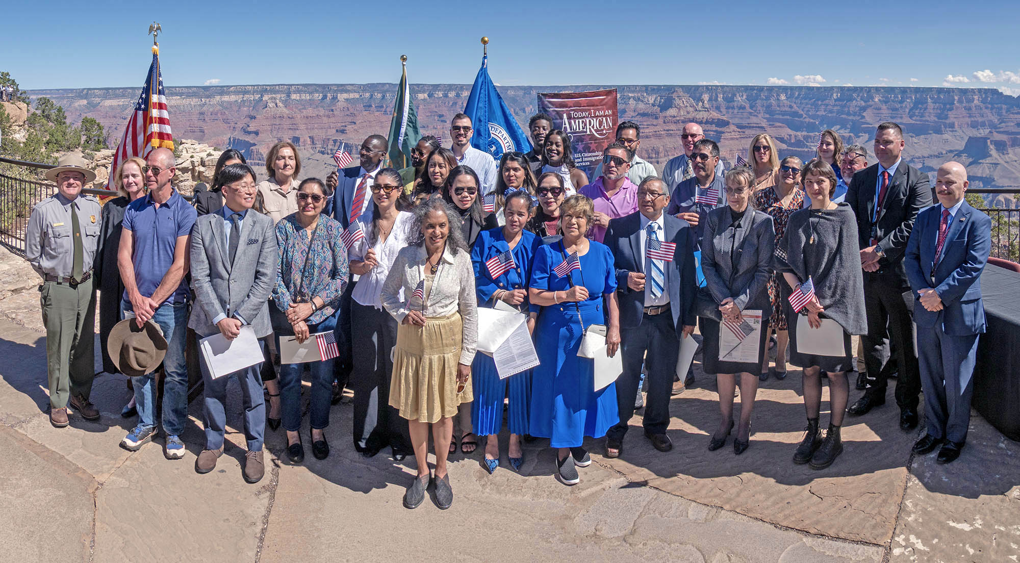 29 new U.S. Citizens are posing for a group photo, holding their papers and small American Flags, with Grand Canyon in the background.
