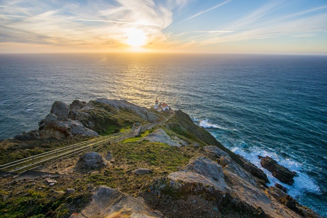 Point Reyes Lighthouse from the Observation Deck at Sunset.  A strip of rocky land juts into the ocean.  A setting sun is seen on the horizon.