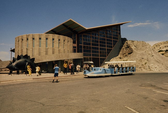 A photo taken from the parking lot shows a building with a tall, glass window walls, an angular roof, and an attached cyclorama