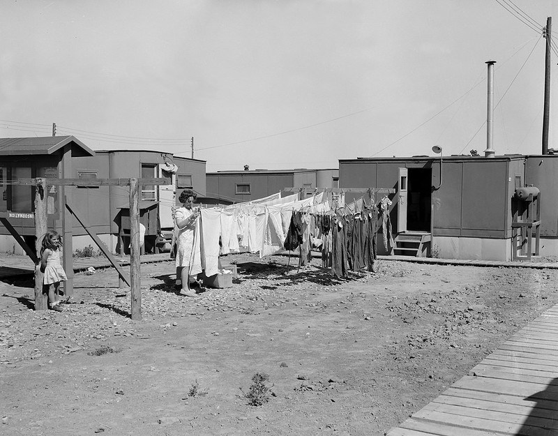 Black and white photo of woman hanging laundry in front of small one- story trailers