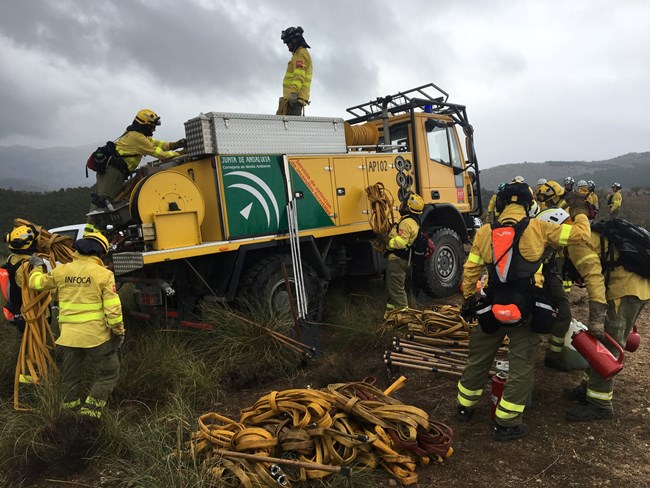 Wildland firefighters in Spain surround a fire engine putting away fire gear.  Fire hose is rolled up on the ground and tools are scattered around the fire engine.