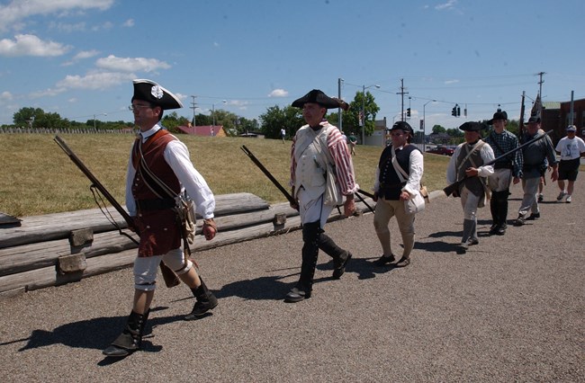 76963265_10156755824931367_8245971453200039936_o Men in 18th C clothing walk in a single file into fort, muskets carried at their right sides.