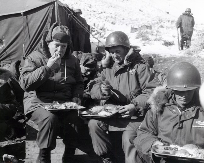 A black and white image showing General Eisenhower wearing a heavy winter parka sitting outdoors eating lunch with other soldiers wearing winter gear