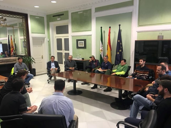 Several people sit around a conference table in an office setting. Flags are on post against a wall in the center.