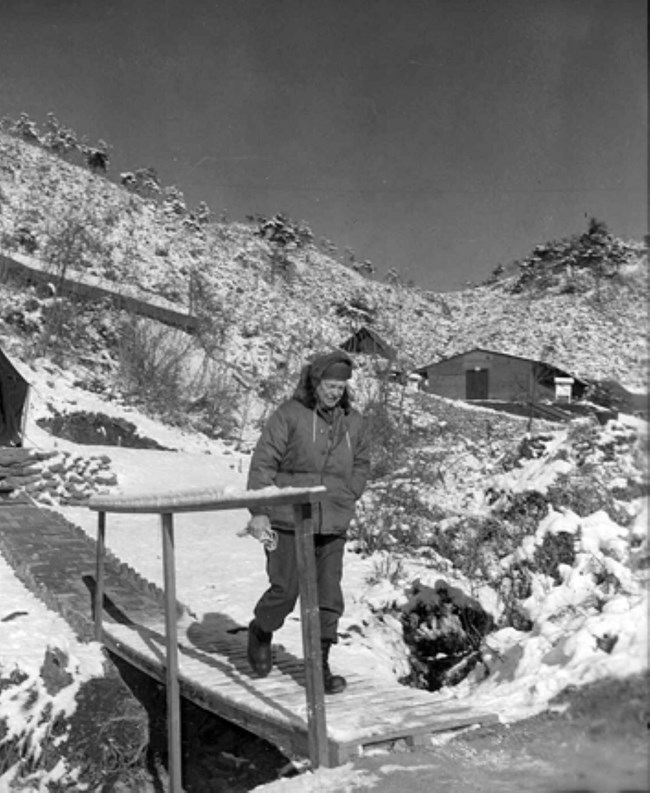 A black and white image of President-Elect Eisenhower wearing a winter coat and hat and walking in a snow covered landscape in Korea