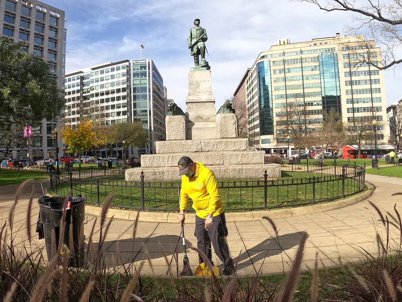 A man in yellow sweeps trash with a statue in the background.