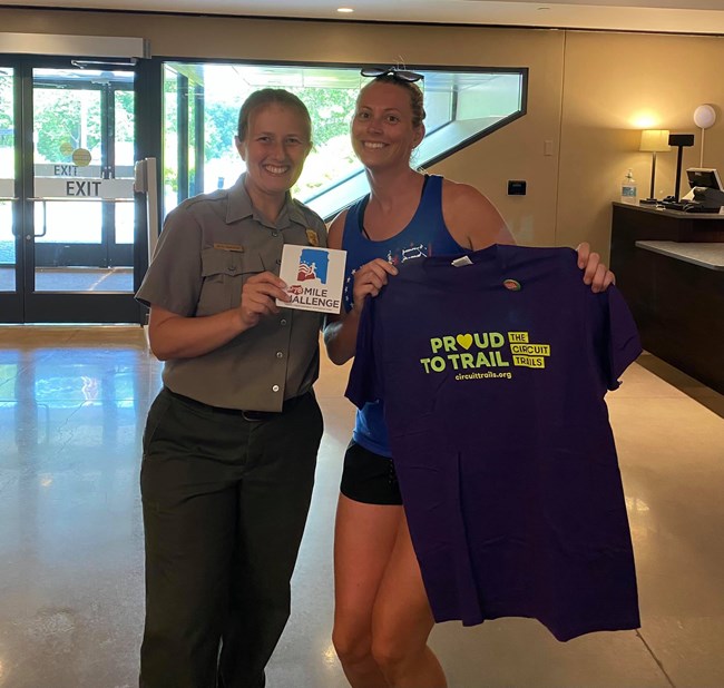78 Mile Challenge Finisher Kimberly a uniformed park ranger and a woman in athletic clothes smile and hold a t-shirt and a magnet