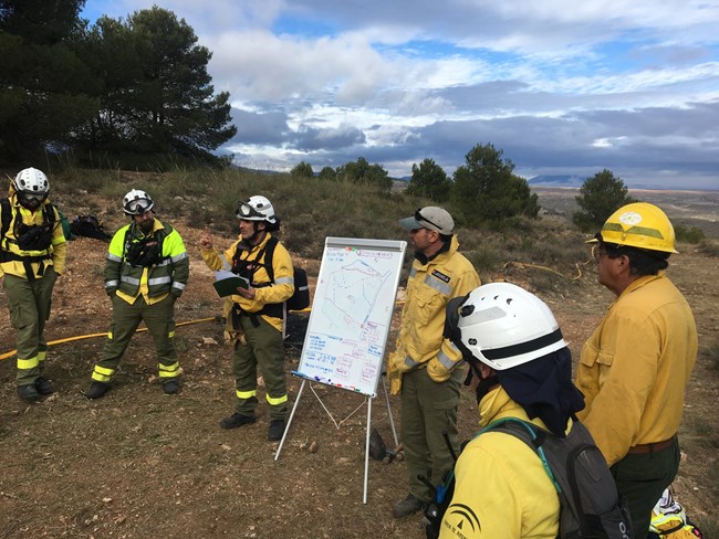 Wildland firefighters from the United States and Spain stand outside by trees in a semi-circle around a white board with a map and words drawn on it.