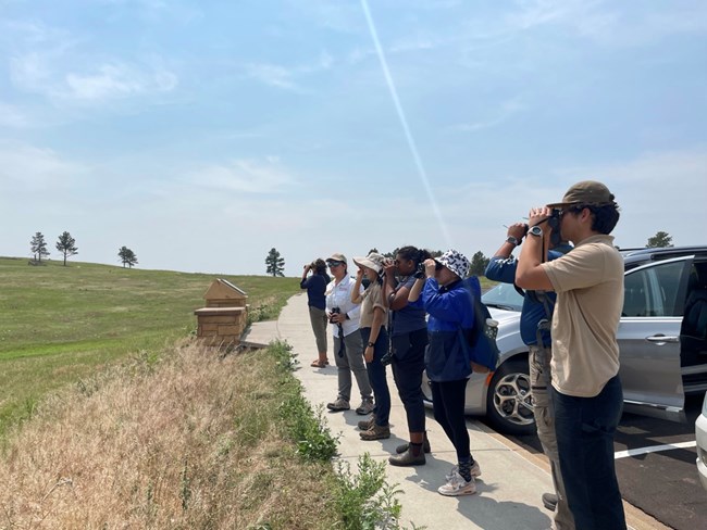 group of people lined up, using their binoculars while looking ahead towards a prairie.