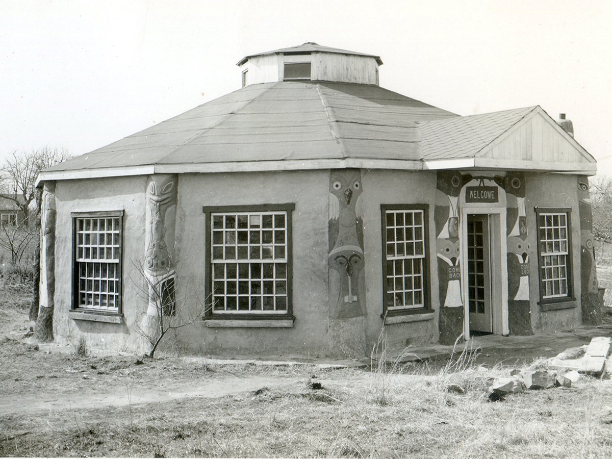 Hexagon shaped building with sloped roof and copula, a large window one each side. Welcome sign over door.