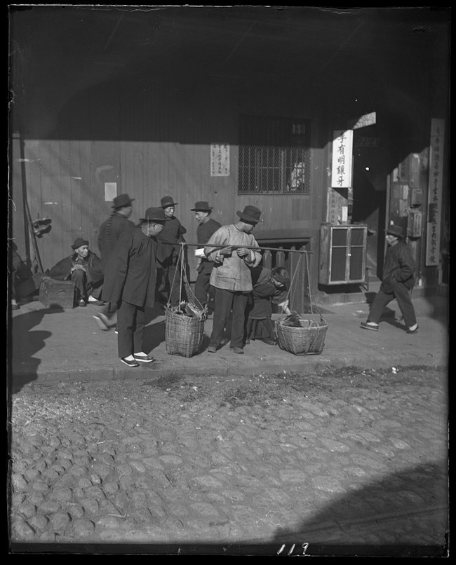 A Chinese immigrant stands outside a wooden building, holding two baskets on a large stick, filled with vegetables.