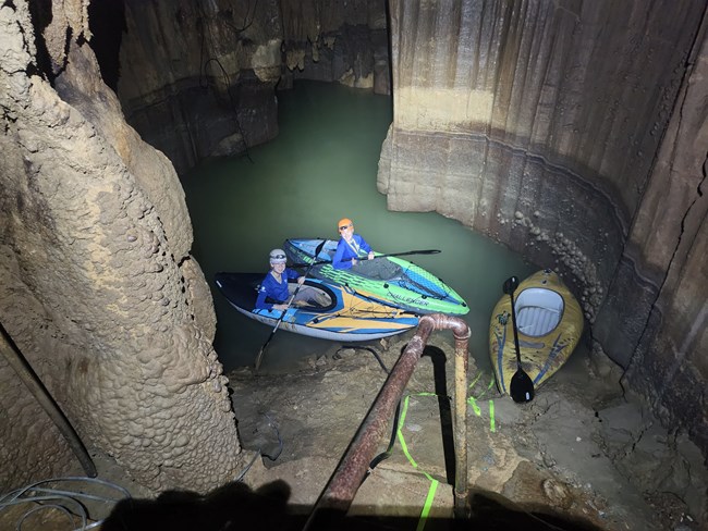 Two people wearing helmets sit in brightly colored inflatable kayaks in a small green body of water inside a cave.