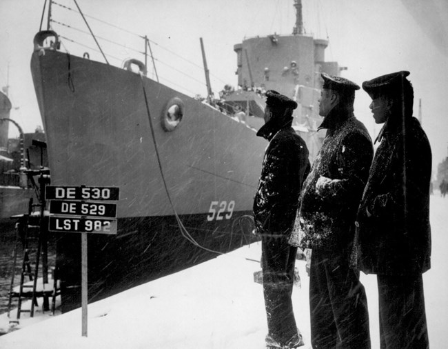 Three African American sailors standing on a pier in the snow beside a destroyer escort.