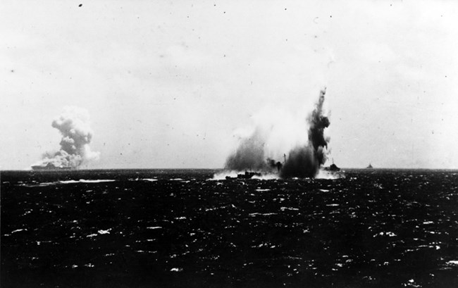 Ship struck by a torpedo in the middle of the ocean with smoke billowing out of it. Another ship with smoke is in the far distance.