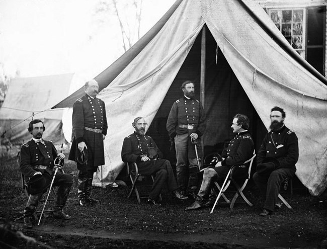 6 men in uniform stand or sit in front of a canvas tent. Hunt leans against his sword. He has a full, trimmed beard and mustache.